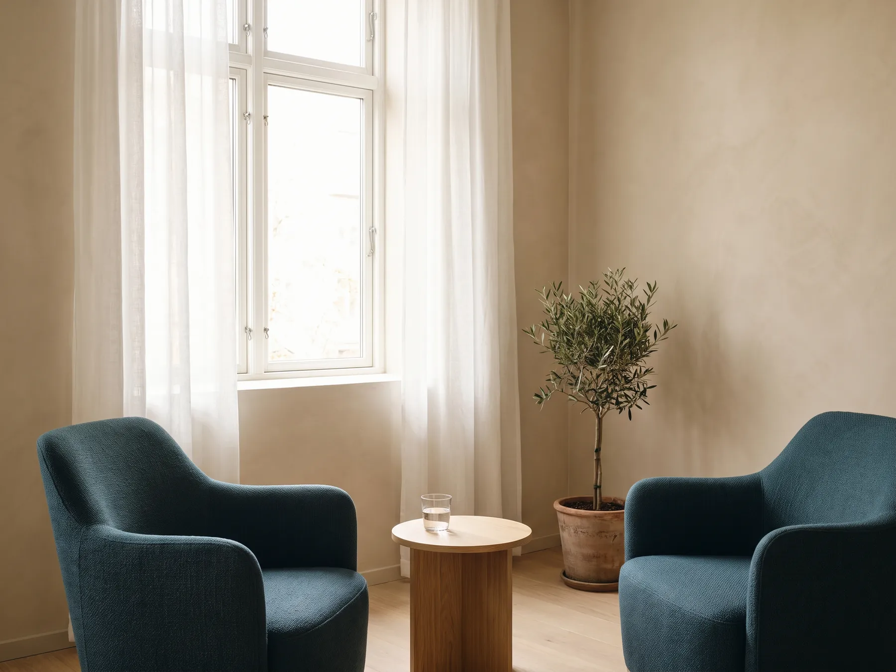 Zwei petrolfarbene Sessel an einem kleinen Holztisch in einem hellen Raum mit Fenster und Olivenbaum